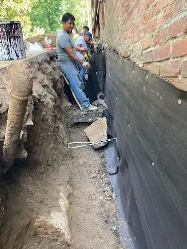 Crew installing black foundation waterproofing membrane on a brick wall in a trench
